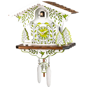  Image of Engstler Quartz Chalet Cuckoo Clock, featuring a white base with a brown roof and intricate green ornaments. A white bench sits in front, surrounded by trees with green details. The pendulum and weights are white with green accents.