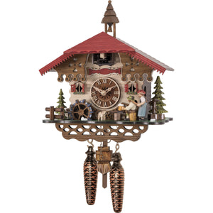  Image of Engstler Quartz Cuckoo Clock depicting a traditional Black Forest scene with a man sitting at a table drinking beer, a woman holding a rolling pin behind him, and a cuckoo bird emerging from a door underneath a red roof with a bell tower.