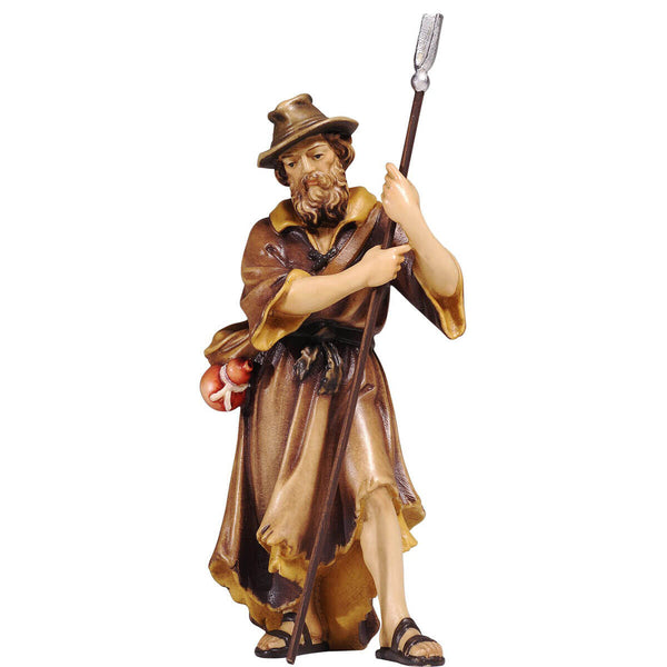 The standing shepherd is dressed in a brown coat. A bottle is strapped to his pouch which is worn as a cross-body bag. The shepherd is wearing a wide-brimmed hat and sandals. He stepped forward with his left leg and he Is leaning on a staff holding it with both hands. He has a beard and his eyes are looking down to the right