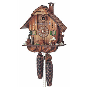 8-Day Black Forest Chalet coocoo clock. A wood chopper is chopping wood on the right where a pile of logs sits and vine is growing up on the side of the dark wood rustic house. A squirrel is eating next to a fence in the grass and an owl is perched on a log on the left. Two windows with green shutters flank the dial and the upper half of the house is painted with flowers. The roof has shingles and a chimney stack.