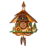 A beautiful traditional chalet-style timepiece with intricate design details. A wood chopper is hard at work to the right, his dog is watching to the left. Trees decorate the corners of the house stained in different brown hues. There is a little balcony on the top floor where the cuckoo bird comes out from behind his door. The bell tower on top of the roof is an additional charming feature.