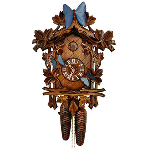 8-Day Traditional Black Forest coocoo clock. The face of the brown wooden clock around the dial is crosshatched. Large carved leaves are growing on a vine around the edge of the clock. There is a blue butterfly on the right lower corner of the clock, another one in the middle on the left side of the clock and a large blue butterfly decorates the highest point of the clock’s crown. The butterflies are moving in sync with the cuckoo bird coming out from the door above the dial on the full and half hour.