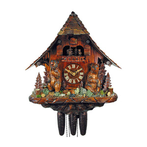  A charming cuckoo clock with a chalet-style design, featuring bears and woodland elements. The clock has a sloping roof with a small door, four dancing bears on the balcony, and two larger bears flanking the clock face. Woodland accents such as brush, logs, and mini fir trees adorn the base, while the sides showcase rustic details like wooden benches, stone walls, and water troughs.