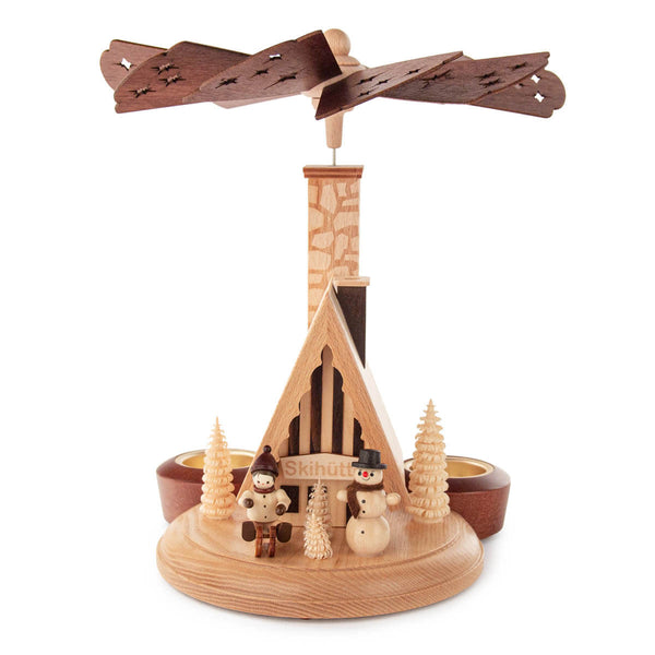 This beautiful, hand carved Christmas pyramid holds a candle on each of the sides of its base to provide gentle lighting on every side when in use. The heat from the candles causes the center of the pyramid to turn, displaying every side of the center design as it rotates. This pyramid features a single tier design with a ski hut – Skihuette and a large chimney. There are several carved trees in natural wood tones, a happy snowman and child dressed for winter fun in front of the hut.