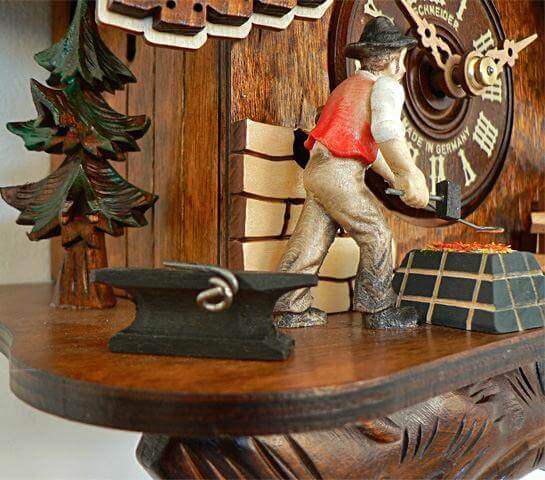 A blacksmith standing between an Anvil and a forge next to the dial of a Schneider cuckoo Clock