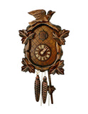 1-Day Schneider Traditional Black Forest Coocoo clock without music. The face of the brown clock is crosshatched. There are four grape leaves with lighter colored carved lines on each of the four corners of the clock. The distinctive feature of this clock is the round clock face. The cuckoo door above the dial opens when the cuckoo bird calls. There is a larger ornately carved cuckoo bird sitting on top of the crown on the roof.