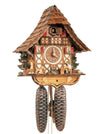 8-Day Black Forest Chalet Schneider coocoo clock with music. The chalet is white with curved brown timbers. A water pump, evergreen tree and stones decorate the left corner while a St. Bernard carrying a small keg watches how a man on a chair taps his beer stein on a table. Behind him on the right is a pile of wood and another fir tree, leaves in the middle. Two windows with red shutters flank the dial. Above is an ornate balcony. The roof is made of shingles.
