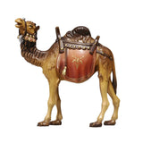 A standing light brown camel with darker mane has turned his head slightly to the left. The mouth is slightly open. The camel is wearing a fine headdress. The dark brown seat sits on top of  a terracotta riding saddle cloth with elaborate decoration. 