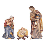 The Holy Family consists of 3 figurines and the manger. Mary, wearing a red dress and blue cape with golden lining and a cream headscarf, is kneeling to the left of Baby Jesus in the manger. Infant Jesus is clad in cloth and lying on a straw lined wooden manger. Baby Jesus is removeable from the manger. Joseph, wearing sandals and dressed in gray with darker cape with golden lining is standing to the right of the manger and is holding a lantern with both hands. 