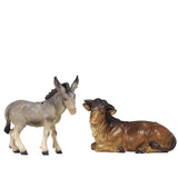 The gray donkey is standing and turns his head slightly to the right. The donkey’s mane is dark. The large brown ox is lying down and has his head turned to the left. Both wooden figurines are carved out of sycamore maple and painted with oil paint.