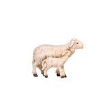 The mother sheep is standing still and looking straight ahead. She has a thick white wool coat. The standing little lamb is cuddling up to his mother on her right side and is leaning his head against her neck looking up to Mom. 