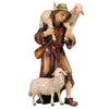 A young shepherd, dressed in a brown coat with belt and a cross-body pouch, is wearing a hat and shoes. His left foot is in front of the other and he is carrying one sheep on his shoulders holding the legs with each hand. A second sheep is standing by the shepherd’s feet looking to the right.