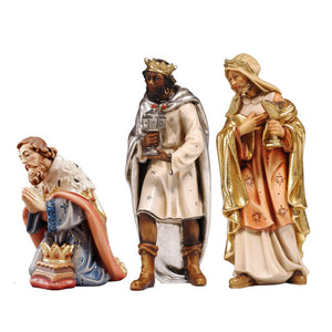 Kneeling King Caspar wears a white-blue gown decorated with fleur-de-lis accents. His cape is terracotta colored with a white ermine cloak, his crown is resting on a stool in front of him, his hands are in prayer. Black standing King Balthazar wears a white gown with a long silver cape and boots. He is holding a chalice with both hands. Standing King Melchior is dressed in a cream and peach gown with golden cape and headscarf, holding his chalice in the left hand, his right hand on his heart. 