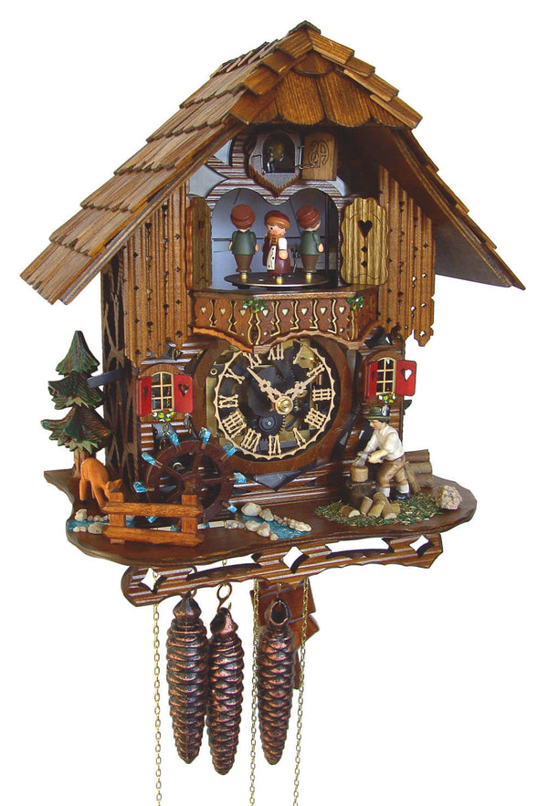 1-Day Schneider Black Forest Chalet Coocoo clock with music. A man is chopping wood on the right side of the platform. On the left, a water wheel spins, and a little brook lined with pebbles on each side is flowing across. There is an evergreen tree and a grazing deer on the left. The clear faced dial shows the sophisticated mechanical movement inside the clock box. Dancers spin on the balcony, above it is the cuckoo clock door underneath the roof with wooden shingles.