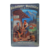 Metal sign of vintage advertisement for Wiedemann's Soft Cheese. An Alpine hiker is seated at a table in front of a wooden house, as a woman serves him a tray of cheese. Two cows rest in the background, in front of an Alpine panorama.
